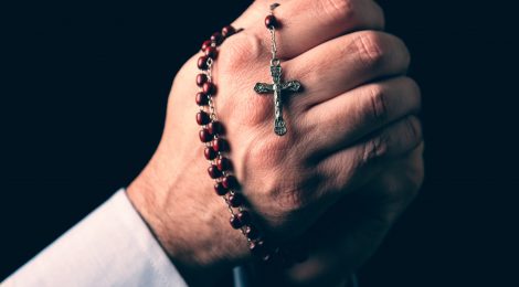 Male hands praying holding a rosary with Jesus Christ in the cross or Crucifix on black background. Mature man with Christian Catholic religious faith
